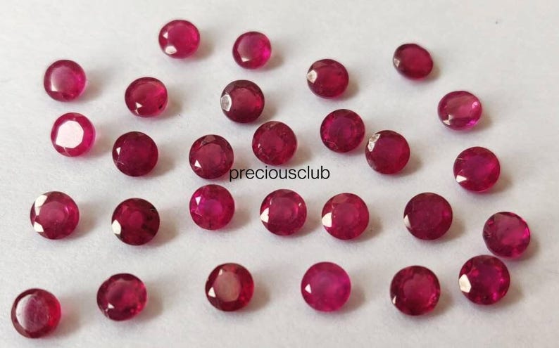 May include: A collection of small, round, faceted red rubies scattered across a plain surface. Each gemstone features a brilliant cut with multiple reflective surfaces. The text "preciousclub" is printed in black in the center of the arrangement.