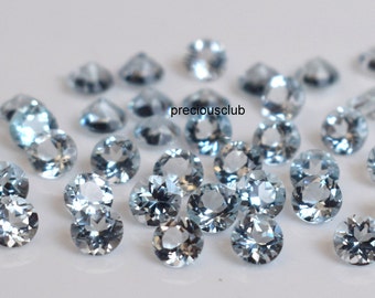 May include: A collection of round, light blue gemstones. The gemstones are faceted and have a clear, glassy appearance. The gemstones are scattered on a white surface. preciousclub