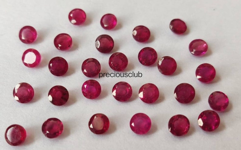 May include: A collection of small, round, faceted red rubies scattered across a white surface. Each gemstone displays a brilliant crimson hue with multiple facets reflecting light. The text "preciousclub" is printed in black in the center.