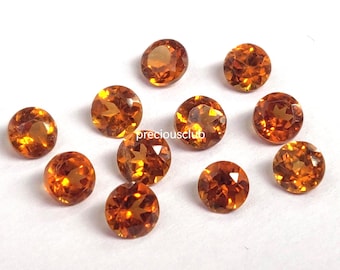 May include: A collection of eleven round, faceted gemstones in shades of orange and amber. The gems are scattered on a white surface, each displaying internal light reflections. The overall effect is one of warmth and brilliance.