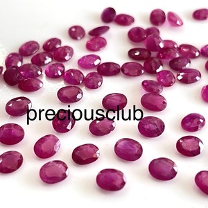 May include: A collection of oval-shaped, faceted ruby gemstones in various sizes, scattered on a white surface. The rubies are a deep, rich red color.