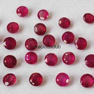 May include: A collection of small, round, faceted red rubies scattered across a plain white surface. Each gemstone displays a brilliant crimson hue with multiple cut surfaces that reflect light. The text preciousclub is printed in black.