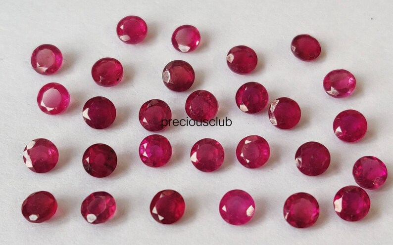 May include: A collection of small, round, faceted red rubies scattered across a plain white surface. Each gemstone displays a brilliant crimson hue with multiple cut surfaces that reflect light. The text preciousclub is printed in black.