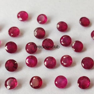 May include: A collection of small, round, faceted rubies with a deep red color and a brilliant, reflective surface. The gemstones are arranged in a loose, scattered pattern on a plain white background. The text preciousclub is visible.