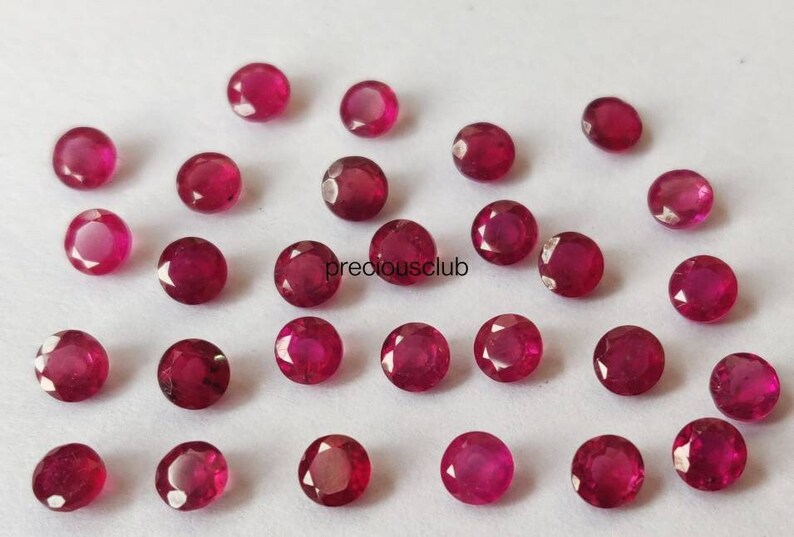 May include: A collection of small, round, faceted rubies with a deep red color and a brilliant, reflective surface. The gemstones are arranged in a loose, scattered pattern on a plain white background. The text preciousclub is visible.