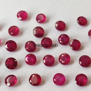 May include: A collection of small, round, faceted rubies with a deep red color and a brilliant, reflective surface. The gemstones are arranged in a loose, scattered pattern on a plain white background. The text preciousclub is visible.
