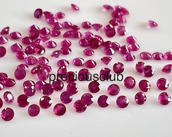 May include: A collection of loose, round, red ruby gemstones. The gemstones are various sizes and are scattered on a white surface.