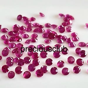 May include: A collection of numerous small, round, faceted magenta-colored gemstones scattered across a plain surface. The text "preciousclub" is printed in black in the center of the arrangement of these loose, polished, vibrant pinkish-red stones.