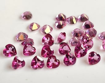 May include: A collection of faceted pink gemstones, some heart-shaped and others with a rounded, multi-faceted design. The gems are scattered across a white surface, catching the light and displaying a range of pink hues.