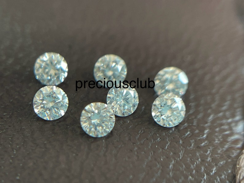 Pode incluir: Sete pedras preciosas redondas e facetadas transparentes, provavelmente diamantes, sobre uma superf&iacute;cie texturizada cinza escura. As pedras preciosas est&atilde;o espalhadas aleatoriamente.