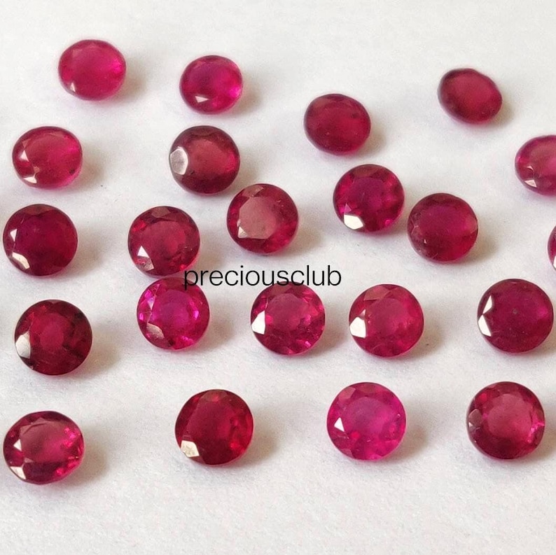 May include: A collection of small, round, faceted red gemstones arranged in a scattered pattern. Each stone features a brilliant cut with multiple reflective surfaces. The text "preciousclub" is printed in black in the center of the image.