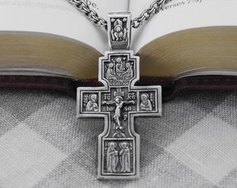 May include: A silver Orthodox cross pendant with intricate details and multiple figures. The cross is on a chain and resting on a book with text.
