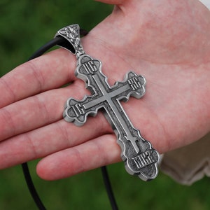 May include: A silver pendant in the shape of a cross with intricate details and inscriptions. The cross is hanging from a black cord.