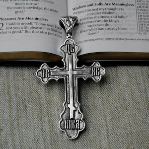 May include: A silver Orthodox cross pendant with intricate details and Cyrillic lettering. The cross is resting on a fabric surface with a Bible open in the background.