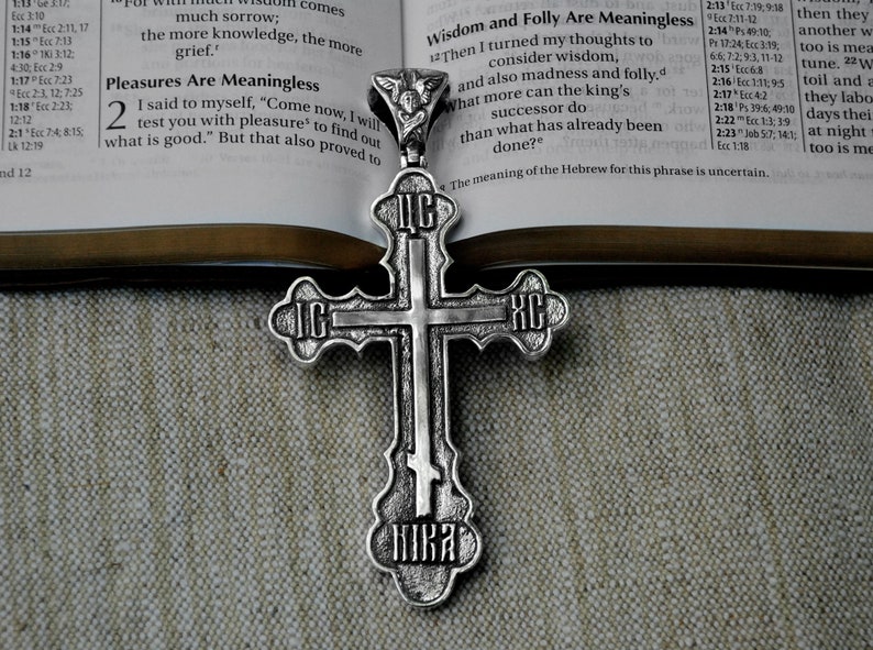May include: A silver Orthodox cross pendant with intricate details and Cyrillic lettering. The cross is resting on a fabric surface with a Bible open in the background.