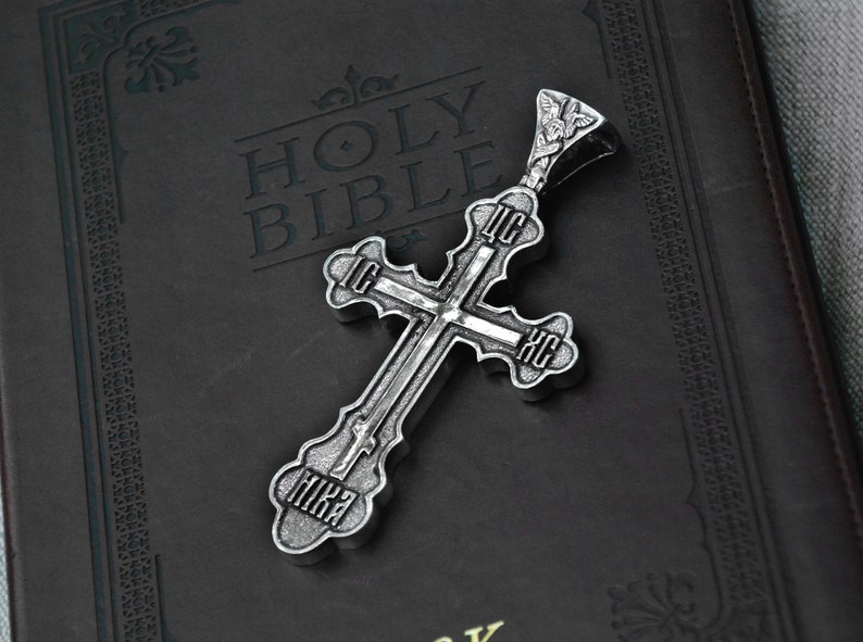 May include: A silver cross pendant with intricate details and Cyrillic lettering. The cross is lying on a black leather-bound Holy Bible.