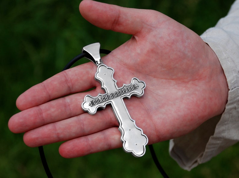 May include: A silver cross pendant with a black cord. The cross has an inscription in Cyrillic script that reads "Спаси и сохрани".