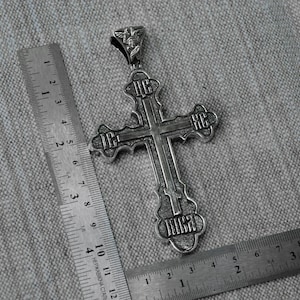 May include: A silver cross pendant with intricate details and engraved lettering. The cross is approximately 2 inches in length and features a small angel figure at the top.