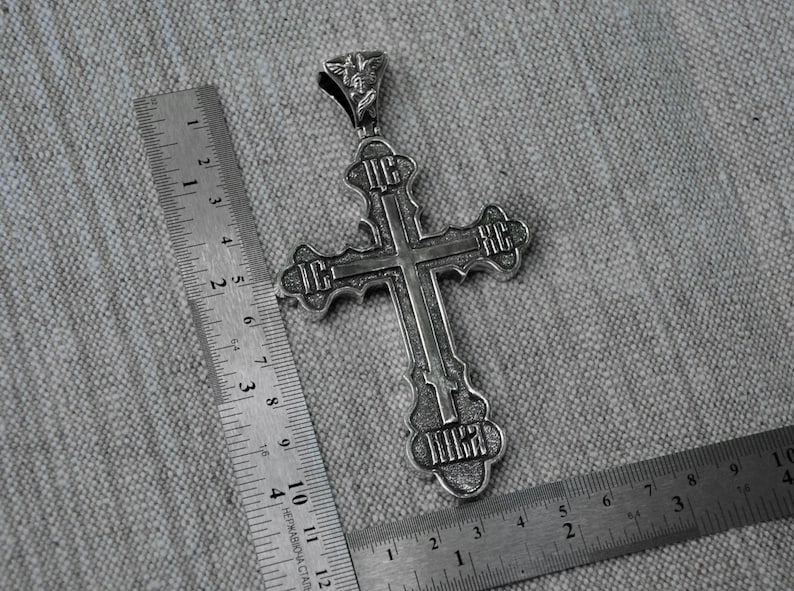 May include: A silver cross pendant with intricate details and engraved lettering. The cross is approximately 2 inches in length and features a small angel figure at the top.