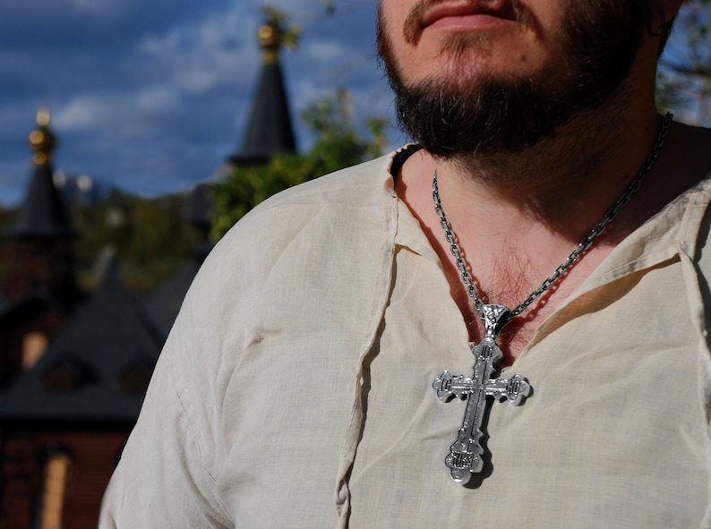 May include: A man wearing a white shirt with a silver chain and a large silver cross pendant.