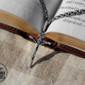 Pode incluir: Um pingente de prata em forma de crucifixo com uma corrente, descansando sobre um livro fechado com uma capa de couro marrom. O pingente apresenta uma figura de Jesus Cristo na cruz.