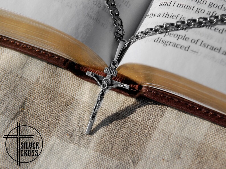 Pode incluir: Um pingente de prata em forma de crucifixo com uma corrente, descansando sobre um livro fechado com uma capa de couro marrom. O pingente apresenta uma figura de Jesus Cristo na cruz.
