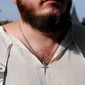 Pode incluir: Um homem vestindo uma camisa de linho branca com um colar de corrente de prata e um pingente de cruz de prata.
