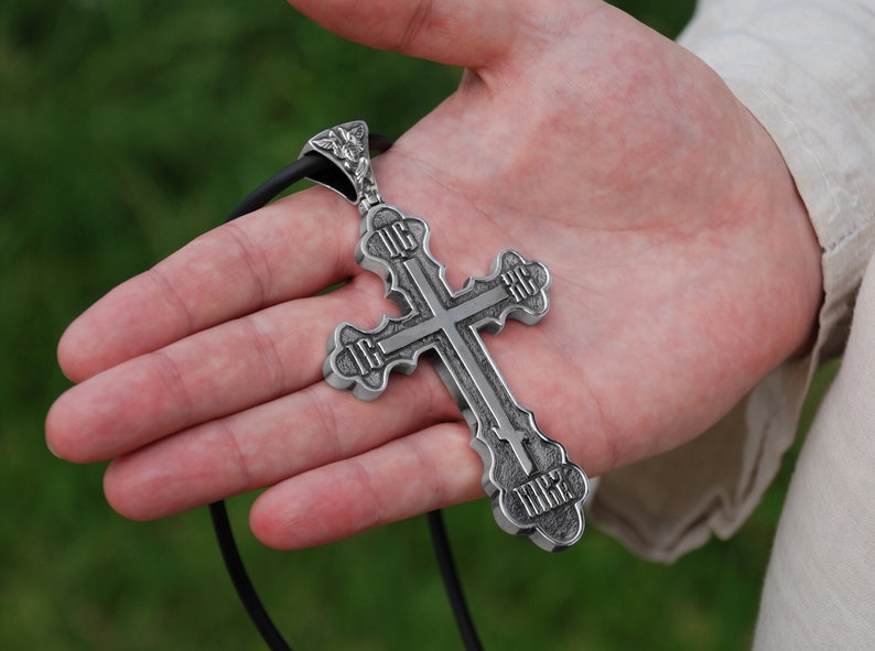 May include: A silver pendant in the shape of a cross with intricate details and inscriptions. The cross is hanging from a black cord.