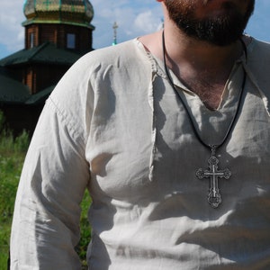 May include: A man wearing a white linen shirt with a black cord necklace and a silver cross pendant.