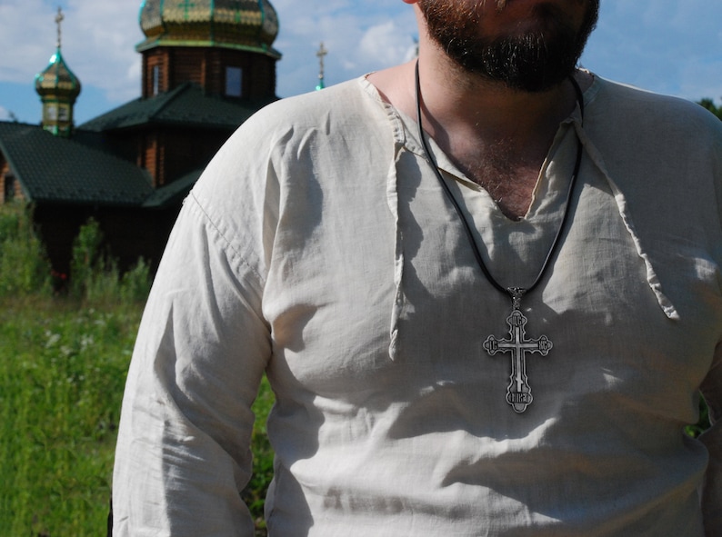 May include: A man wearing a white linen shirt with a black cord necklace and a silver cross pendant.