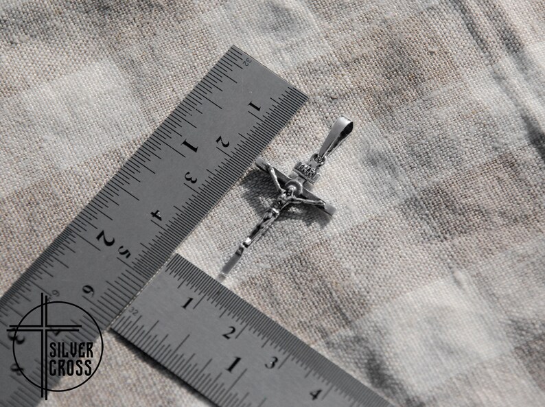 Pode incluir: Um pingente de prata em forma de crucifixo com uma figura detalhada de Jesus Cristo. O pingente &eacute; medido com uma r&eacute;gua, mostrando que tem aproximadamente 5 cm de comprimento.