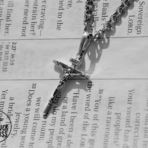 Pode incluir: Um colar de corrente de prata com um pingente de crucifixo de prata. O crucifixo apresenta uma figura de Jesus Cristo na cruz. O colar est&aacute; sobre uma p&aacute;gina de um livro com texto.