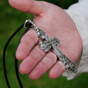 May include: A silver cross pendant with intricate details hangs from a black leather cord. The cross is held in a person's hand.