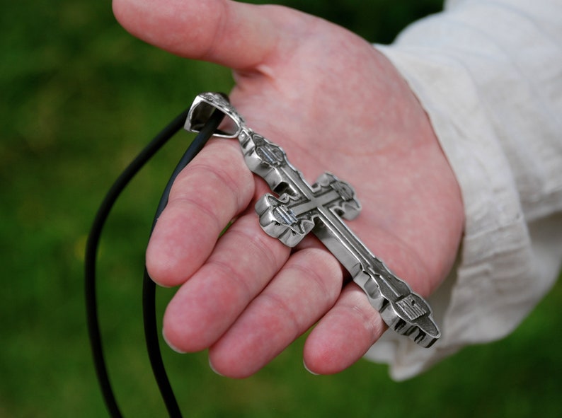 May include: A silver cross pendant with intricate details hangs from a black leather cord. The cross is held in a person's hand.
