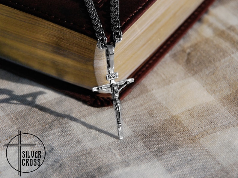 Pode incluir: Um colar de corrente de prata com um pingente de crucifixo de prata. O crucifixo apresenta uma representa&ccedil;&atilde;o detalhada de Jesus Cristo. O colar est&aacute; sobre um livro encadernado em couro castanho.