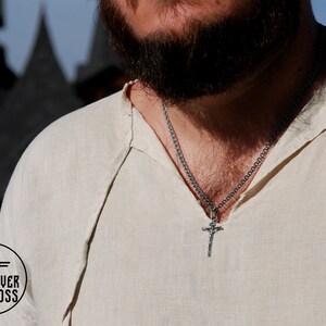 Pode incluir: Um homem vestindo uma camisa de linho branca com um colar de corrente de prata e um pingente de cruz de prata.