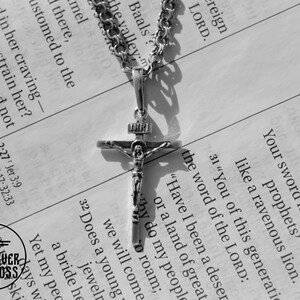 Pode incluir: Um colar de corrente de prata com um pingente de crucifixo de prata. O pingente apresenta uma figura de Jesus Cristo na cruz. O colar est&aacute; sobre uma p&aacute;gina de um livro com texto sobre a palavra do Senhor.