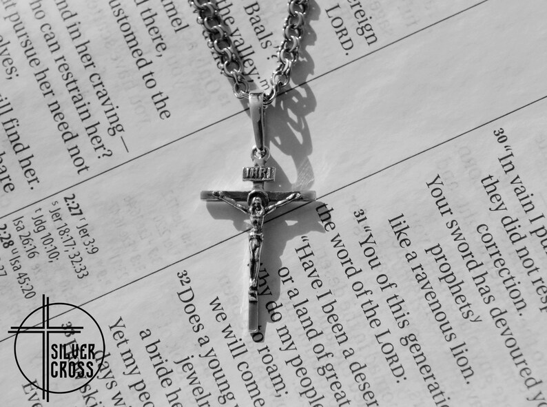 Pode incluir: Um colar de corrente de prata com um pingente de crucifixo de prata. O pingente apresenta uma figura de Jesus Cristo na cruz. O colar est&aacute; sobre uma p&aacute;gina de um livro com texto sobre a palavra do Senhor.