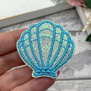 Op de afbeelding: Een schelpvormige patch met een glinsterend wit interieur en een turquoise geborduurde omtrek. De patch heeft een gedetailleerd ontwerp met een geschulpte rand en is waarschijnlijk voor decoratieve doeleinden.