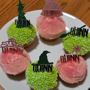 May include: Six cupcakes on a white plate. Three have pink frosting, and three have green frosting with sprinkles. Each cupcake is topped with a glittery black topper that spells "Quinn". The toppers are shaped like a witch's hat, a princess, and a crown.