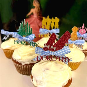 May include: Cupcakes with white frosting and decorative toppers. Toppers include ruby red slippers, a yellow silhouette of characters, a green castle, and a pink crown. Each cupcake has a blue and white gingham bow and the name "Dorothy".