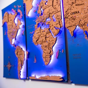 Peut inclure: Une carte du monde en trois panneaux en liège, montée sur un fond bleu. Les continents sont bruns, avec un éclairage LED bleu soulignant les bords. Comprend une petite découpe d'avion. La carte est un objet décoratif sur le thème du voyage.