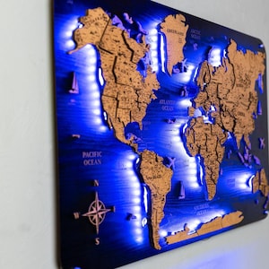 以下が含まれることがあります： 青い背景とLEDライトで照らされた木製の世界地図。大陸は薄茶色で、海は濃い青色です。羅針盤が左下にあります。
