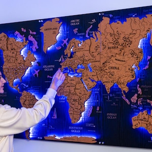 Peut inclure: Une grande carte du monde en bois avec un fond bleu foncé et des continents bruns. La carte est éclairée par des lumières LED bleues, mettant en évidence les océans. La personne pointe la carte. La carte comprend le texte "GREENLAND", "ATLANTIC OCEAN", "ARCTIC OCEAN", "RUSSIA", "CHINA", "PACIFIC OCEAN", "INDIAN OCEAN", et "SOUTHERN OCEAN".