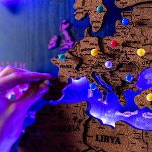 A hand is placing a blue pushpin on a cork world map, which is illuminated with blue light.