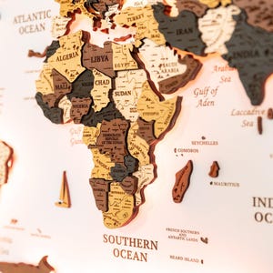 May include: A wooden world map with detailed country outlines in various shades of brown and grey. The map includes labels for oceans, seas, and countries, with a focus on the African continent. The map is mounted on a white surface.