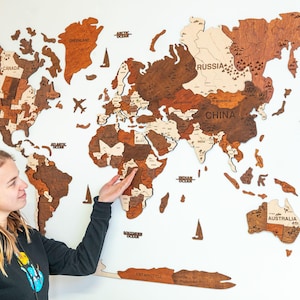 以下が含まれることがあります： 白壁に設置された、茶色とベージュの色合いの木製世界地図。 地図には国名と、飛行機とヨットの装飾的な切り抜きが含まれています。 人が地図を指しています。