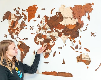 May include: A wooden world map with countries in varying shades of brown and tan is mounted on a white wall. The map includes country names and decorative airplane and sailboat cutouts. A person is pointing at the map.
