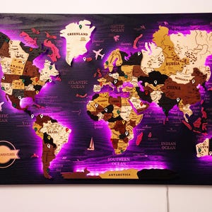 Può includere: Mappa del mondo in legno con illuminazione a LED viola. La mappa presenta vari paesi in diverse tonalità di legno, con oceani in legno scuro. Include icone di aereo e barca a vela. Il testo "OUR ADVENTURES" è visibile.
