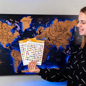 A woman is holding a map of the world with various country flags and icons on it, standing in front of a wall with a large world map.